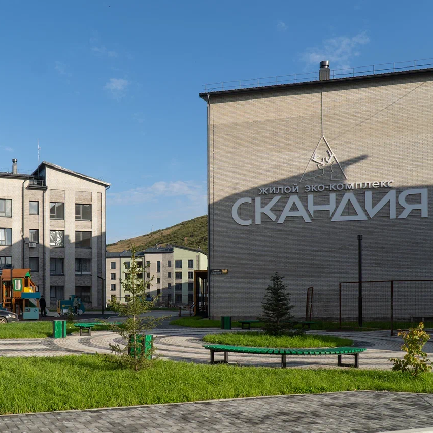 Жилой комплекс «Скандия» с благоустроенной территорией. Видны современные многоэтажные дома, детская игровая площадка, зеленые газоны, скамейки и пешеходные дорожки.  Небо ясное, солнечное.