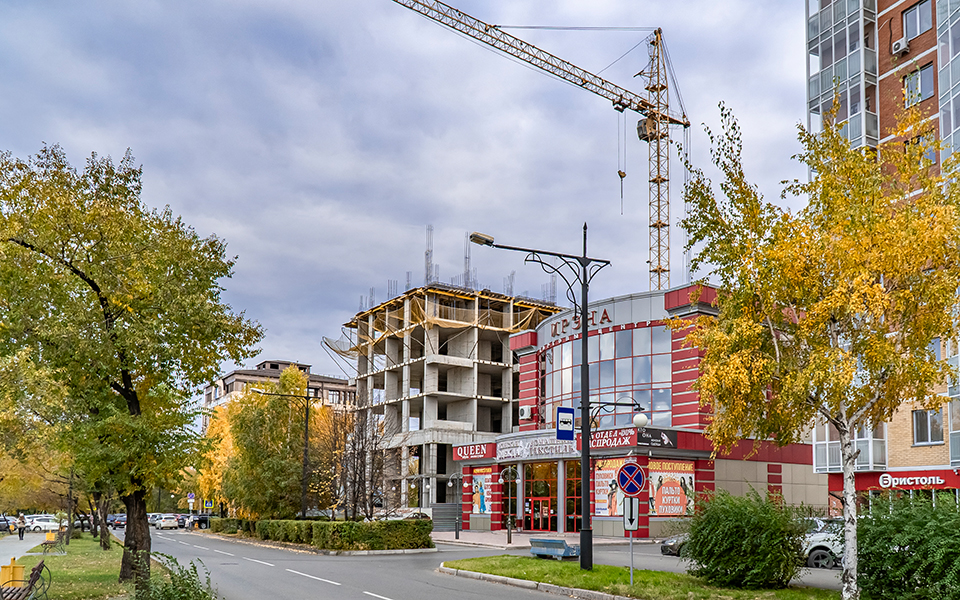 Улица в осеннем городе с зданиями, одним из которых является строящийся объект с башенным краном, и деревьями с желтой листвой.